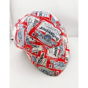 May include: A multi-panel cap featuring an all-over print of the Budweiser logo in red, white, and blue. The fabric displays repeated text and graphics including the word Budweiser, the phrase King of Beers, and the word Genuine in a repeating pattern.