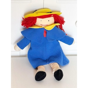 May include: A handmade doll with red yarn hair, a yellow hat, and a blue coat. The doll has a white face with simple facial features and is wearing a yellow collar. The doll's legs are tan with black shoes.