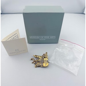 Könnte beinhalten: Goldfarbene Katzen- und Geigen-Ohrring-Anstecknadel vom Museum of Fine Arts, Boston. Die Anstecknadel zeigt eine Katze, die Geige spielt. Die Anstecknadel wird mit der Originalverpackung und einer Informationskarte gezeigt.
