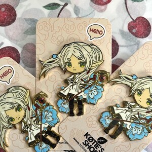 May include: Enamel pins depicting a blonde anime-style character with green eyes, wearing a white dress with blue floral accents. Each pin is on a tan card with the text "Hello" and "Kotie's Shop". The background is a cherry pattern.