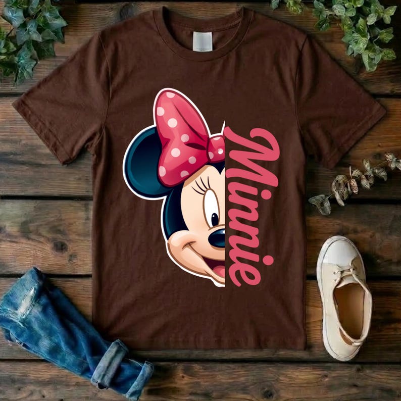 May include: A brown short-sleeved t-shirt featuring a graphic of half of Minnie Mouse's face with a pink polka-dot bow and the name Minnie written in large pink script lettering vertically along the right side of the character illustration.