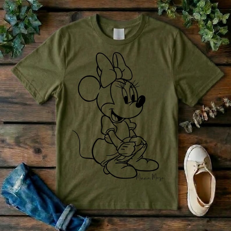 May include: An olive green short-sleeved t-shirt featuring a black line art graphic of Minnie Mouse on the front. The character is depicted in a seated pose with her signature bow. The shirt has a crew neckline and a standard fit.