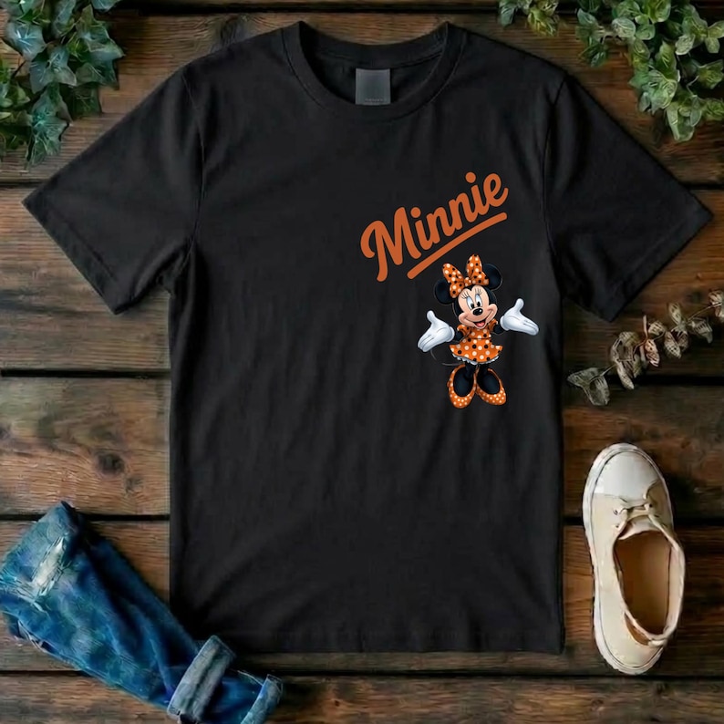 Puede incluir: Una camiseta negra de manga corta con el nombre Minnie escrito en letras naranjas sobre un gr&aacute;fico del personaje Minnie Mouse, que lleva un vestido de lunares naranja y negro con guantes blancos y zapatos a juego en naranja y negro.