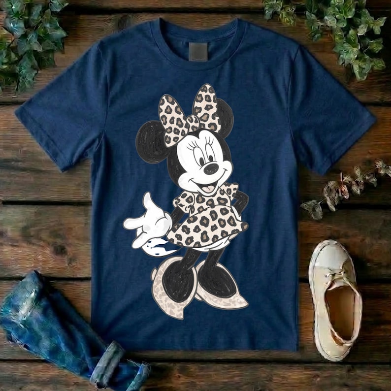 Op de afbeelding: Een marineblauw t-shirt met korte mouwen en een afbeelding van een tekenfilmmuis. Het personage draagt een jurk en een strik, beide versierd met een beige en zwart luipaardpatroon. De muis heeft witte handschoenen en lichtgekleurde schoenen.