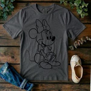 May include: A charcoal gray short-sleeve t-shirt featuring a black line art graphic of Minnie Mouse sitting down. The character is depicted with her signature bow and dress. The script text Minnie Mouse is printed in black near the bottom right.