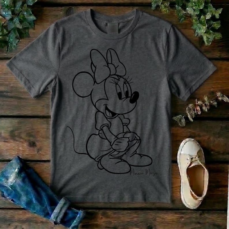 May include: A charcoal gray short-sleeve t-shirt featuring a black line art graphic of Minnie Mouse sitting down. The character is depicted with her signature bow and dress. The script text Minnie Mouse is printed in black near the bottom right.