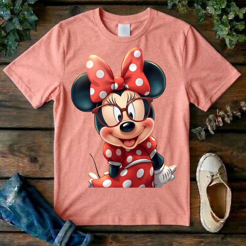 May include: A peach-colored short-sleeve t-shirt featuring a graphic of a cartoon mouse wearing large brown-rimmed glasses and a red dress with white polka dots. The mouse also wears a matching red bow with white polka dots on its head.