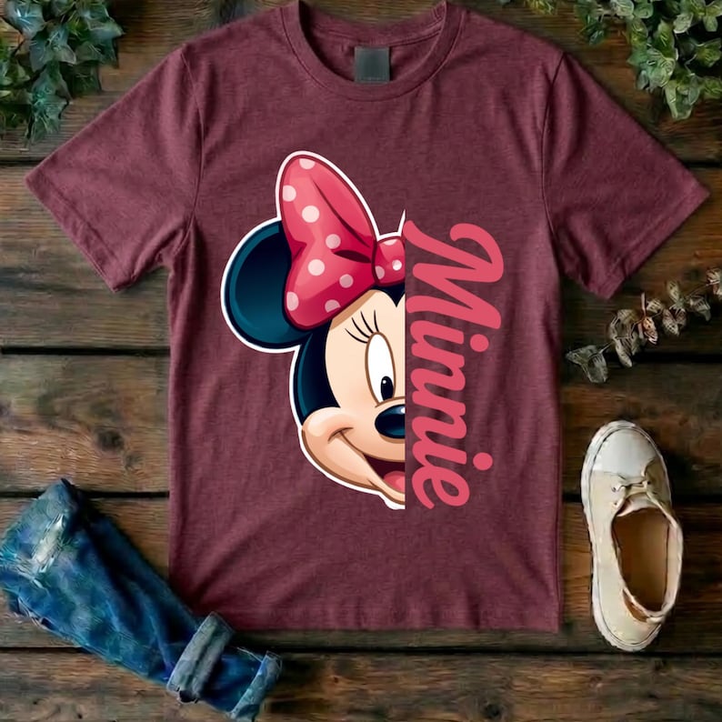 May include: A maroon short-sleeved t-shirt featuring a graphic of half of Minnie Mouse's face with a pink and white polka-dot bow. The word Minnie is printed in a large, vertical, pink script font along the right side of the graphic design.