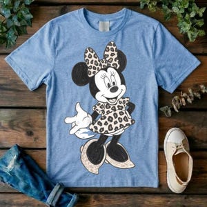 Op de afbeelding: Een lichtblauw T-shirt met korte mouwen en een afbeelding van een tekenfilmmuis. Het personage draagt een jurk en een strik, beide versierd met een bruin en zwart luipaardpatroon. De muis heeft zwarte oren, witte handschoenen en lichte schoenen.