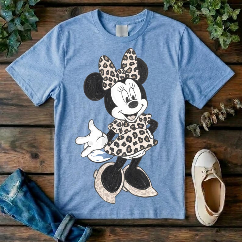 Op de afbeelding: Een lichtblauw T-shirt met korte mouwen en een afbeelding van een tekenfilmmuis. Het personage draagt een jurk en een strik, beide versierd met een bruin en zwart luipaardpatroon. De muis heeft zwarte oren, witte handschoenen en lichte schoenen.