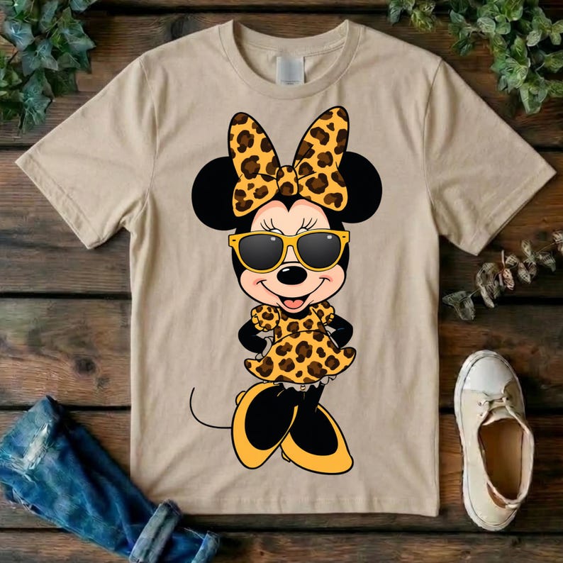 May include: Tan t-shirt featuring a cartoon Minnie Mouse character wearing sunglasses and a leopard print bow, dress, and shoes. The character has black ears and a smiling face. The shirt is on a wooden surface.