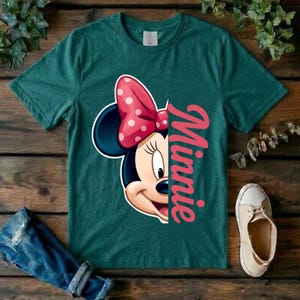 May include: A dark teal short-sleeve t-shirt featuring a graphic of half of Minnie Mouse's face with a pink polka-dot bow. The name Minnie is printed vertically in large pink script letters next to the character illustration on the front.