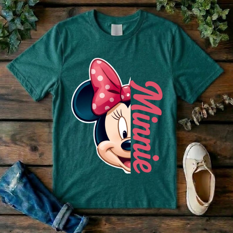 May include: A dark teal short-sleeve t-shirt featuring a graphic of half of Minnie Mouse's face with a pink polka-dot bow. The name Minnie is printed vertically in large pink script letters next to the character illustration on the front.