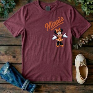 Puede incluir: Una camiseta de manga corta color granate con un gr&aacute;fico de Minnie Mouse con un vestido de lunares y un lazo naranja, con la palabra Minnie impresa en letra cursiva naranja sobre el personaje. La camiseta tiene cuello redondo y corte est&aacute;ndar.