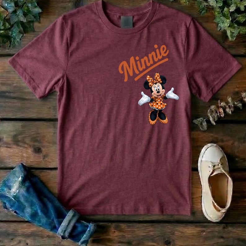 Puede incluir: Una camiseta de manga corta color granate con un gr&aacute;fico de Minnie Mouse con un vestido de lunares y un lazo naranja, con la palabra Minnie impresa en letra cursiva naranja sobre el personaje. La camiseta tiene cuello redondo y corte est&aacute;ndar.