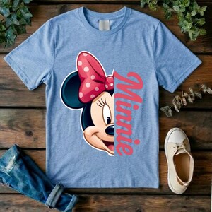 May include: A light blue short-sleeve t-shirt featuring a graphic of the right side of Minnie Mouse's face, including a pink bow with white polka dots, positioned next to the vertical pink script text that reads Minnie.