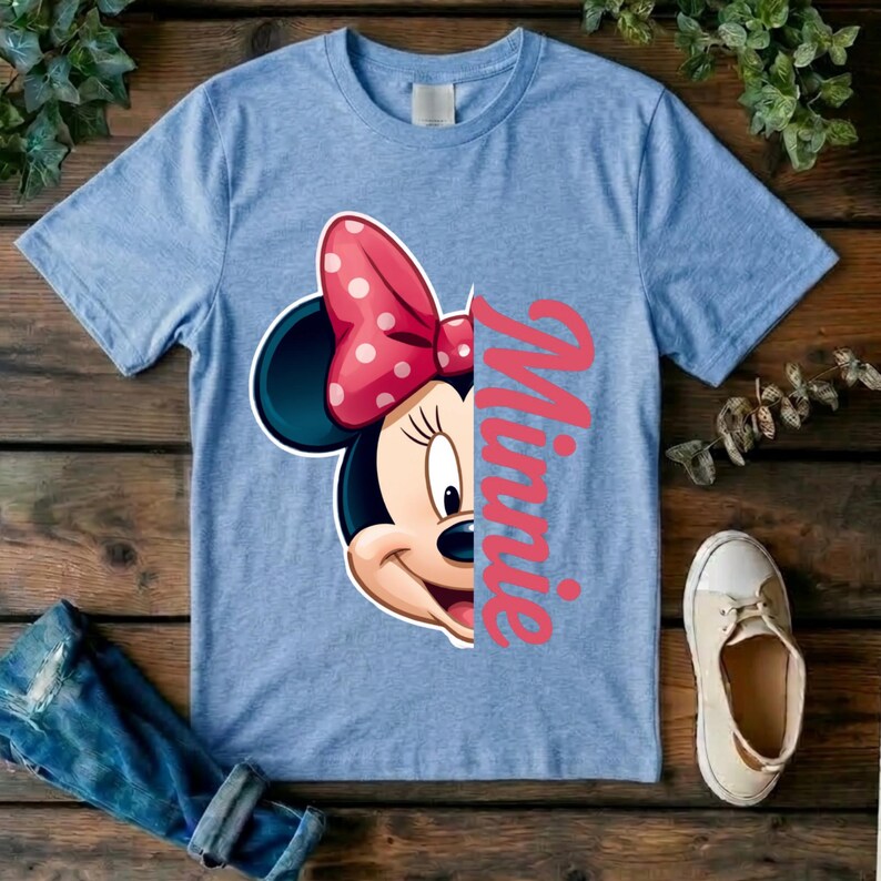 May include: A light blue short-sleeve t-shirt featuring a graphic of the right side of Minnie Mouse's face, including a pink bow with white polka dots, positioned next to the vertical pink script text that reads Minnie.