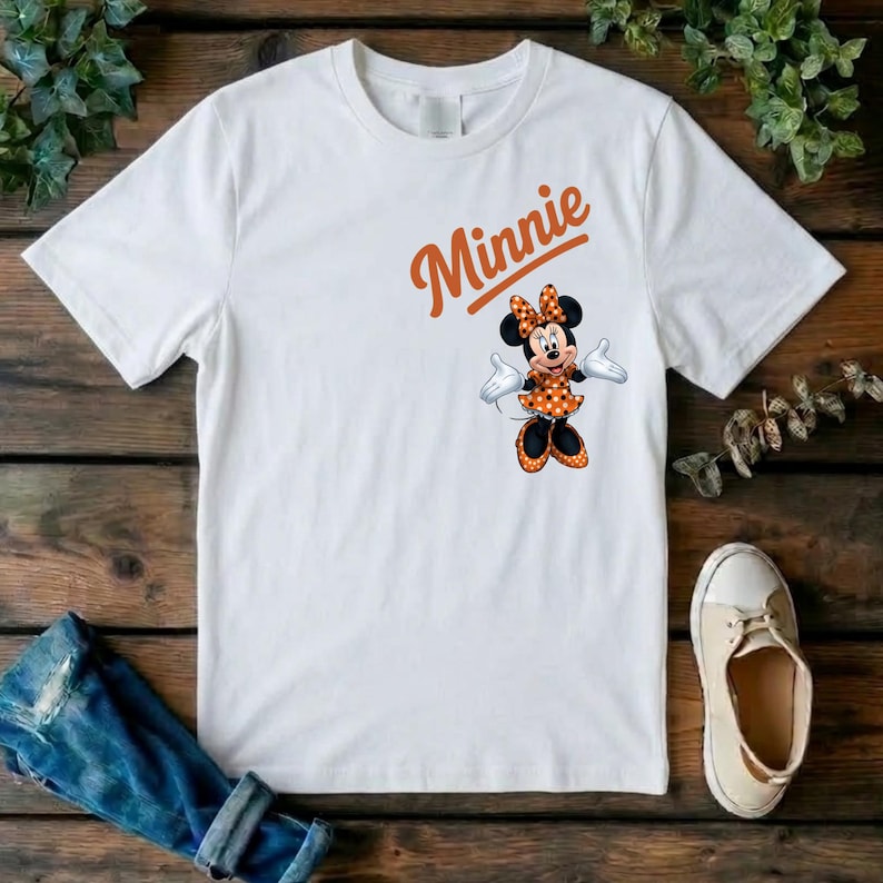 Puede incluir: Una camiseta blanca de manga corta con un gr&aacute;fico de Minnie Mouse con un vestido y un lazo de lunares naranjas y blancos. El nombre Minnie est&aacute; impreso en letra cursiva naranja sobre el personaje, situado en la parte superior derecha del pecho.