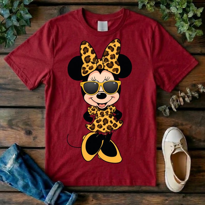 May include: A maroon t-shirt featuring a cartoon Minnie Mouse character wearing sunglasses, a bow, and a dress, all with a leopard print design. The character's shoes are yellow and black. The shirt is displayed on a wooden surface.