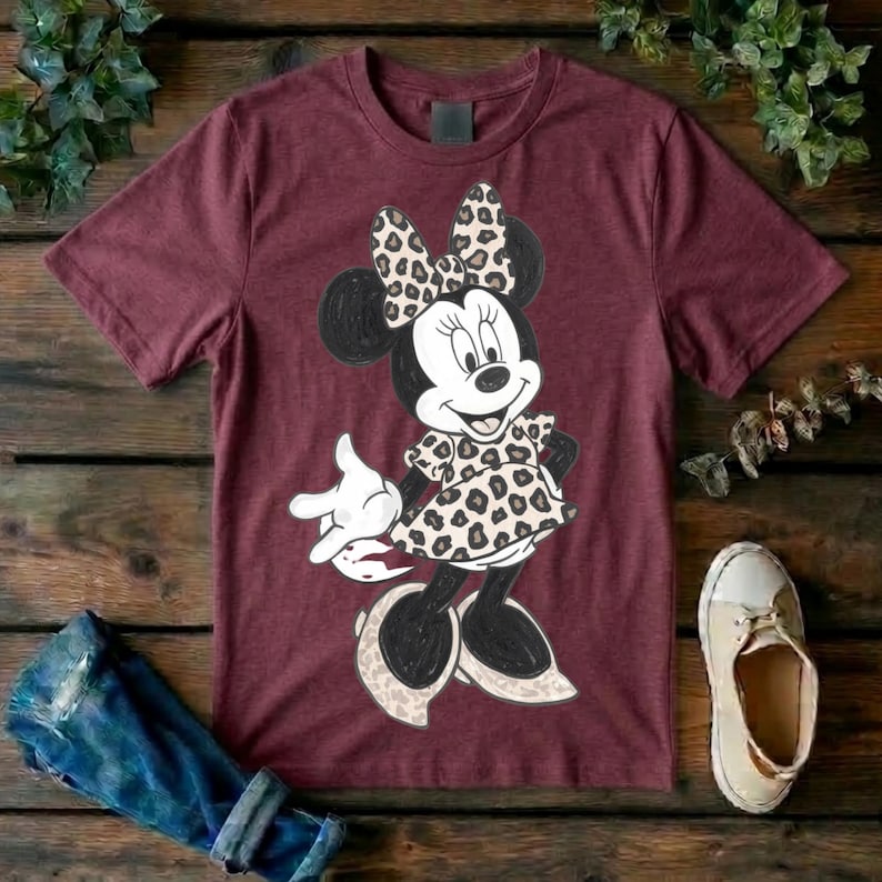 Op de afbeelding: Een bordeauxrood T-shirt met korte mouwen en een afbeelding van een tekenfilmmuis die een strik en een jurk met een beige en zwart luipaardpatroon draagt. Het personage heeft zwarte oren, een wit gezicht en staat in een staande houding.