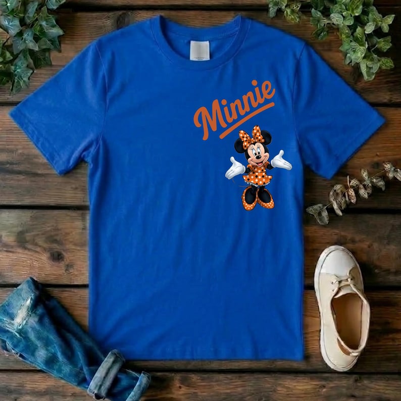Puede incluir: Una camiseta de manga corta de color azul real con el nombre Minnie escrito en letra naranja sobre un gr&aacute;fico del personaje Minnie Mouse, que viste un vestido naranja de lunares con un lazo a juego, guantes blancos y zapatos naranjas.