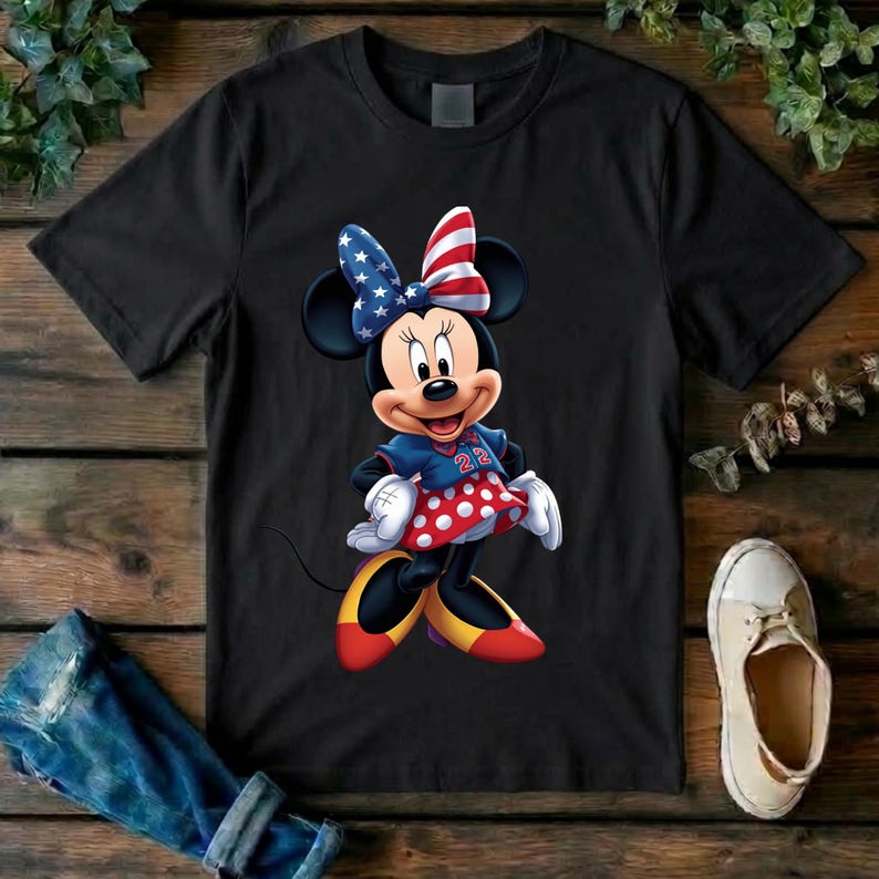 May include: A black short-sleeved t-shirt featuring a graphic of Minnie Mouse wearing a blue jersey with the number 22, a red and white polka dot skirt, yellow shoes, and a bow patterned with the American flag stars and stripes design.