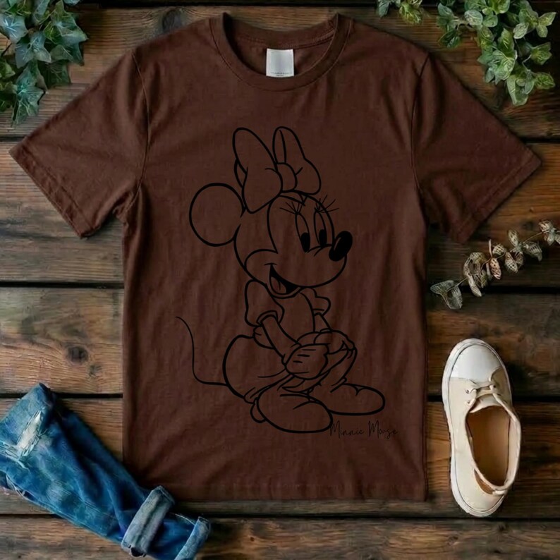 May include: A brown short-sleeved t-shirt featuring a black line art graphic of Minnie Mouse sitting down. The character is depicted with her signature bow and dress. The shirt has a crew neckline and a plain back, with the name Minnie Mouse written in cursive script near the bottom of the graphic.