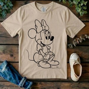May include: A beige short-sleeved t-shirt featuring a black line art illustration of Minnie Mouse on the front. The character is depicted in a seated pose with a large bow on her head. The name Minnie Mouse is written in cursive script below.