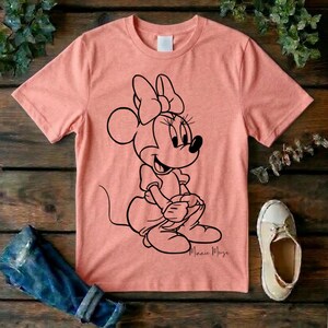 May include: A peach-colored short-sleeve t-shirt featuring a black line art graphic of Minnie Mouse sitting down. The character is depicted with her signature bow and dress. The script text Minnie Mouse is printed in black below the character graphic.