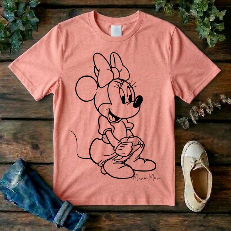 May include: A peach-colored short-sleeve t-shirt featuring a black line art graphic of Minnie Mouse sitting down. The character is depicted with her signature bow and dress. The script text Minnie Mouse is printed in black below the character graphic.