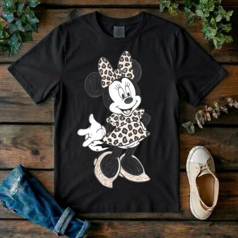 Op de afbeelding: Een zwart t-shirt met korte mouwen en een afbeelding van een tekenfilmmuis. Het personage draagt een jurk en een strik, beide versierd met een beige en zwart luipaardpatroon. Het personage heeft witte handschoenen en schoenen aan.