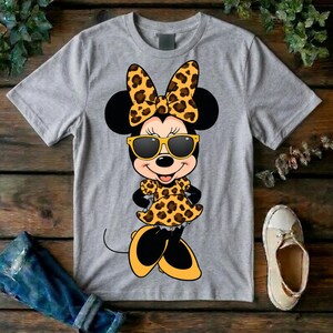 May include: A gray t-shirt featuring a cartoon Minnie Mouse design. Minnie Mouse is wearing a leopard print bow, dress, and sunglasses. Her shoes and the sunglasses frames are yellow. The design is centered on the shirt.