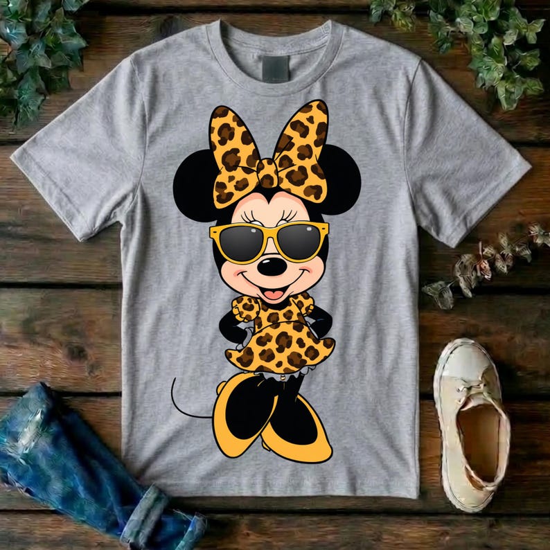 May include: A gray t-shirt featuring a cartoon Minnie Mouse design. Minnie Mouse is wearing a leopard print bow, dress, and sunglasses. Her shoes and the sunglasses frames are yellow. The design is centered on the shirt.