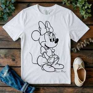 May include: A white short-sleeved t-shirt featuring a black line art graphic of Minnie Mouse sitting down. The character is depicted with her signature bow, dress, and shoes. The text Minnie Mouse is written in a cursive script below the graphic.