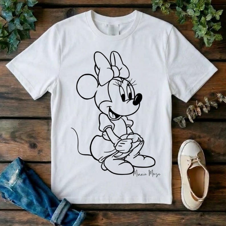 May include: A white short-sleeved t-shirt featuring a black line art graphic of Minnie Mouse sitting down. The character is depicted with her signature bow, dress, and shoes. The text Minnie Mouse is written in a cursive script below the graphic.