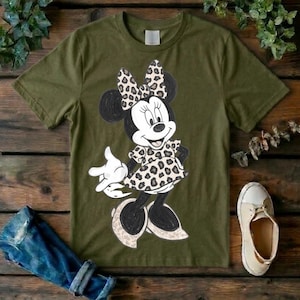 Op de afbeelding: Een olijfgroen T-shirt met korte mouwen en een grafische illustratie van een tekenfilmmuis. Het personage draagt een jurk en een strik, beide versierd met een beige en zwart luipaardpatroon, terwijl de rest van de figuur zwart is.