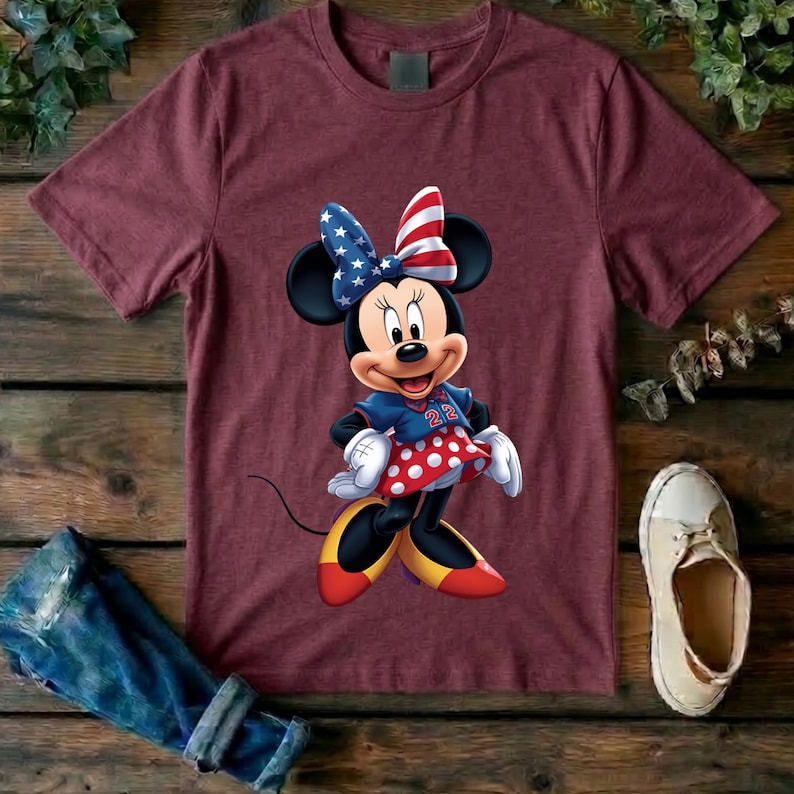 May include: A maroon short-sleeved t-shirt featuring a graphic of a cartoon mouse character wearing a patriotic bow with stars and stripes, a blue top with the number 22, and a red skirt with white polka dots, paired with yellow and red shoes.