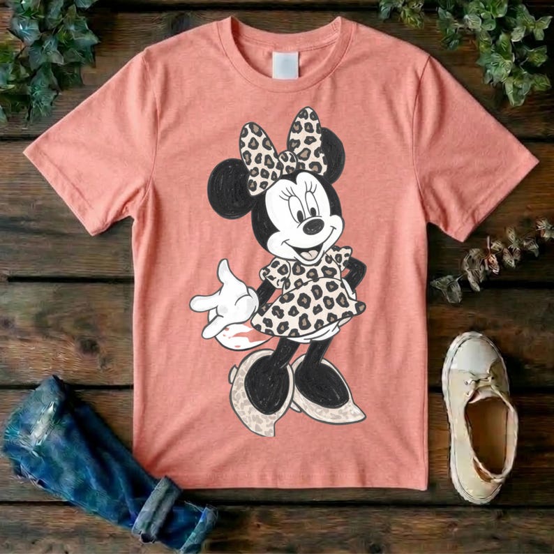 Op de afbeelding: Een perzikkleurig T-shirt met korte mouwen en een afbeelding van een tekenfilmmuis. Het personage draagt een jurk en een strik, beide versierd met een zwart en beige luipaardprint. De muis heeft zwarte oren, armen en benen.