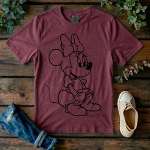 May include: A maroon short-sleeved t-shirt featuring a black line art graphic of Minnie Mouse sitting down. The character is depicted with her signature bow and dress. The shirt has a crew neckline and a heathered texture throughout the fabric.