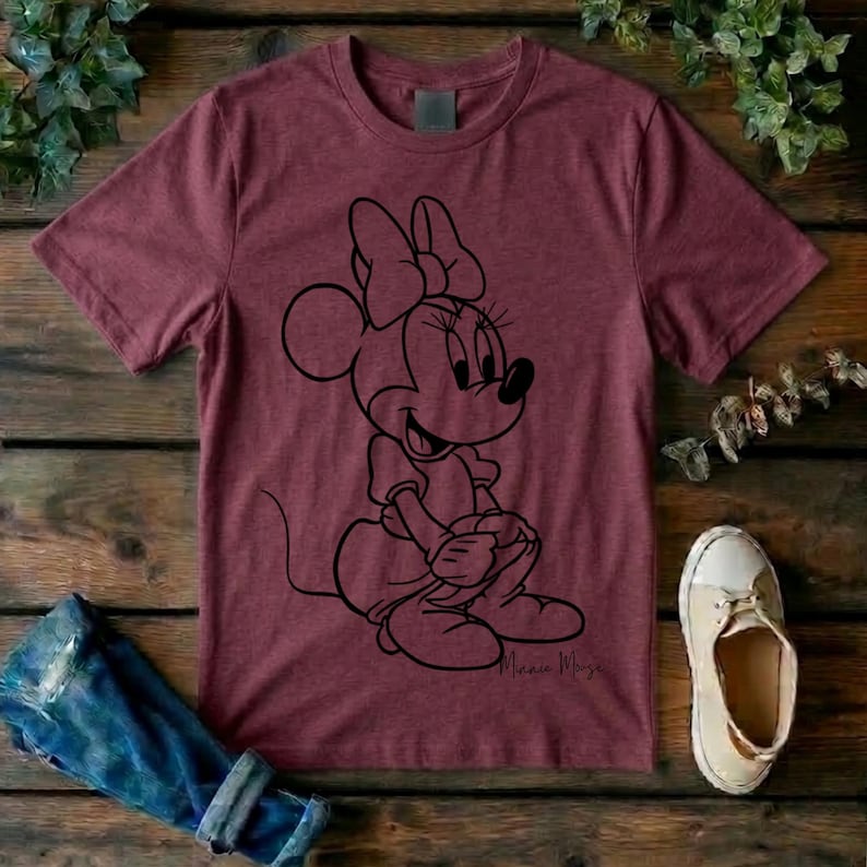 May include: A maroon short-sleeved t-shirt featuring a black line art graphic of Minnie Mouse sitting down. The character is depicted with her signature bow and dress. The shirt has a crew neckline and a heathered texture throughout the fabric.
