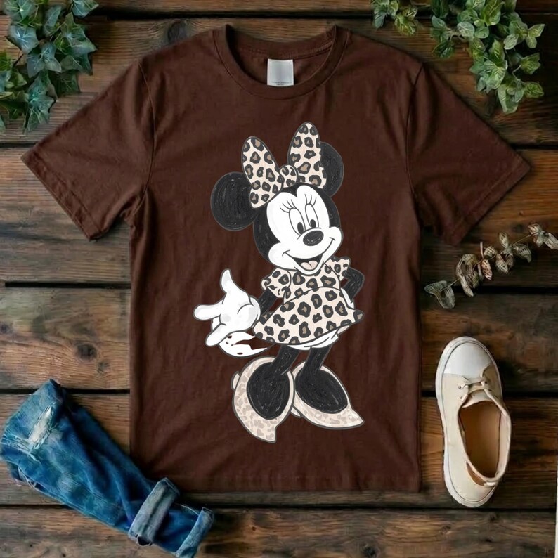 Op de afbeelding: Een bruin T-shirt met korte mouwen en een afbeelding van een cartoonmuisfiguur in een jurk en strik met een beige en zwart luipaardpatroon. Het figuur heeft witte handschoenen en schoenen, met een zwarte omlijning en schetsachtige details.