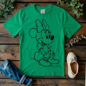 May include: A bright green short-sleeved t-shirt featuring a black line art graphic of Minnie Mouse sitting down. The character is depicted with her signature bow and dress. The shirt has a crew neckline and a plain back, presented on a flat surface.