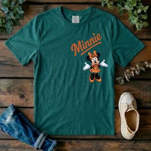 Puede incluir: Una camiseta de manga corta de color verde oscuro con la palabra Minnie en texto naranja sobre un gr&aacute;fico del personaje Minnie Mouse con un vestido y lazo de lunares naranjas y blancos, guantes blancos y zapatos negros.