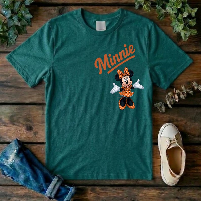 Puede incluir: Una camiseta de manga corta de color verde oscuro con la palabra Minnie en texto naranja sobre un gr&aacute;fico del personaje Minnie Mouse con un vestido y lazo de lunares naranjas y blancos, guantes blancos y zapatos negros.
