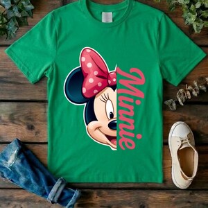 May include: A bright green short-sleeved t-shirt featuring a graphic of half of Minnie Mouse's face, including her signature pink and white polka-dot bow. The word Minnie is printed vertically in large pink script letters next to the character.