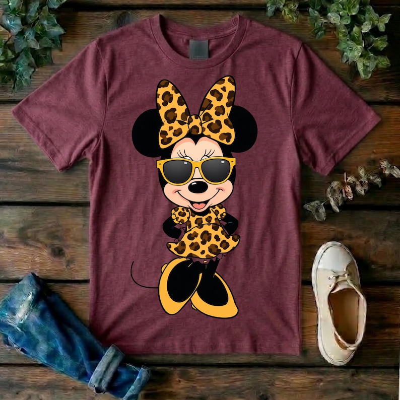 May include: Maroon t-shirt featuring a cartoon Minnie Mouse character wearing sunglasses, a bow, and a dress with a leopard print design. The character's shoes are yellow and black.