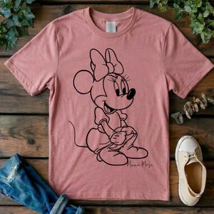 May include: A dusty rose short-sleeved t-shirt featuring a black line art graphic of Minnie Mouse sitting down. The character is depicted with her signature bow and dress. The text "Minnie Mouse" is printed in a cursive script below the graphic.