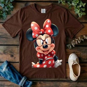 May include: A brown short-sleeved t-shirt featuring a graphic of Minnie Mouse wearing red-rimmed glasses and a matching red dress with white polka dots. The character has a large red bow with white polka dots between her ears and white gloves.