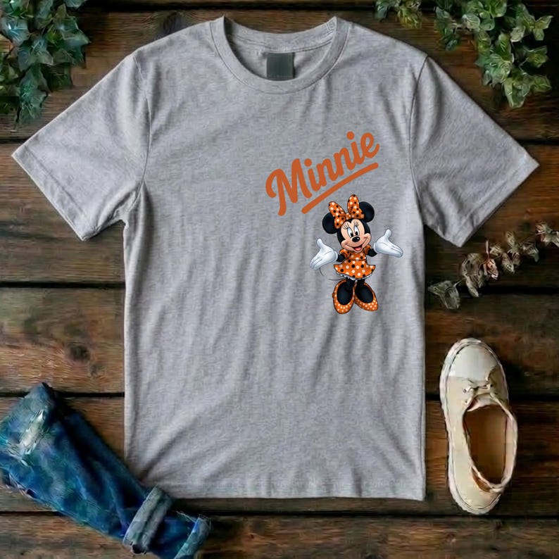 Puede incluir: Una camiseta gris jaspeado de manga corta con el nombre Minnie en texto naranja sobre un gr&aacute;fico del personaje Minnie Mouse vistiendo un vestido de lunares naranja y negro con lazo a juego, guantes blancos y zapatos de color negro.