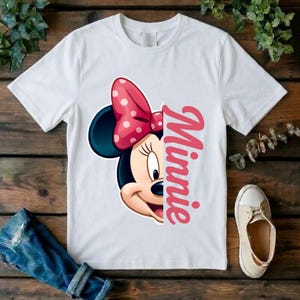 May include: A white short-sleeved t-shirt featuring a graphic of the left half of Minnie Mouse's face, complete with a pink polka-dot bow. To the right of the character graphic, the name Minnie is printed vertically in a bold pink script font.